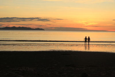 Silhouette children standing at beach against sky during sunset