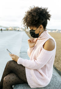 Woman with curly hair wearing face mask using smart phone while sitting on retaining wall