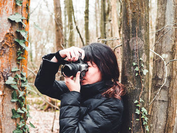 Young woman photographing through tree trunk in forest