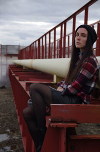 Young woman looking away while sitting on seat against sky