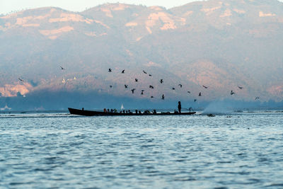 Scenic view of inle lake with silhouette of birds and woman steering boat with people against sky