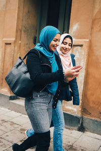 Smiling young woman walking with female friend holding smart phone in city