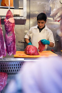 Hispanic male butcher standing behind metal counter and slicing fresh meat during work on market