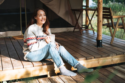 Brunette woman in nordic sweater drinking tea and relaxing in glamping in nature