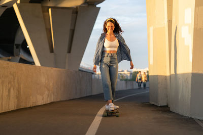 Trendy young girl skate on longboard dressed denim shirt, white top and stylish sunglasses at sunset