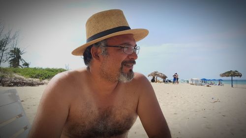 Shirtless man wearing hat and eyeglasses at beach against sky