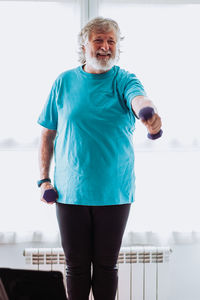 Happy senior bearded male in sportswear smiling and doing exercise with dumbbells during fitness workout in morning at home