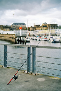 Fishing rod and sailboats moored on harbor by buildings in city against sky