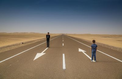 Brothers standing on road arrow symbols along landscape against clear blue sky