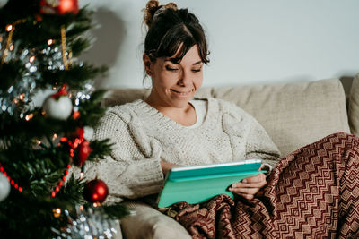 Young woman using digital tablet while sitting on christmas tree