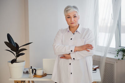 Gray-haired woman white shirt standing at workplace with laptop and planner