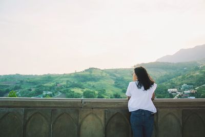 Rear view of woman leaning on retaining wall against clear sky