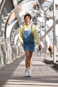 Low angle full body of happy young hispanic female with curly hair wearing denim overall dress with yellow sweatshirt and sneakers walking on enclosed footbridge in city