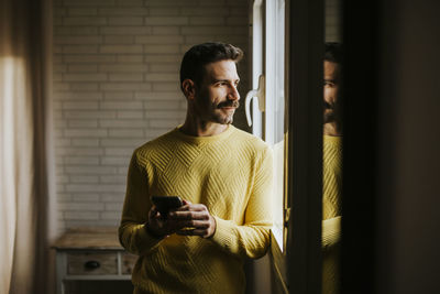 Contemplating man looking through window while holding mobile phone at home