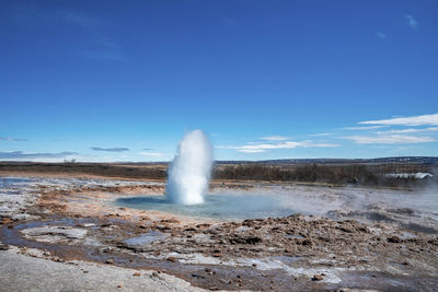 Eruption of strokkur geyser amidst landscape against blue sky during sunny day