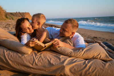 A young couple with a child lies on the seashore and reads a book.