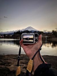 Cropped hand photographing lake with camera against sky