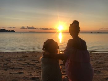 Rear view of woman sitting with dog at beach against orange sky