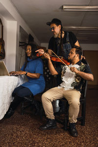 Serious multiracial men in casual outfit learning playing violin near woman at table with laptop in bright apartment
