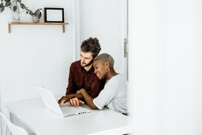 Side view of contemporary african american woman sitting at table and working with laptop with young man surfing digital table at home