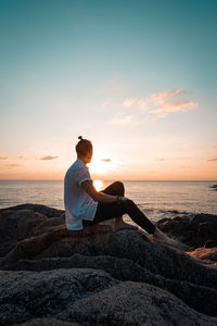 Man sitting on rock looking at sea against sky during sunset