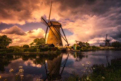 Traditional windmill by lake against cloudy sky during sunset