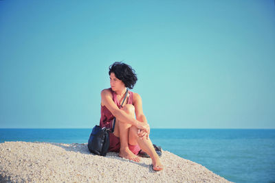 Full length of thoughtful woman looking away while sitting on rock against sea