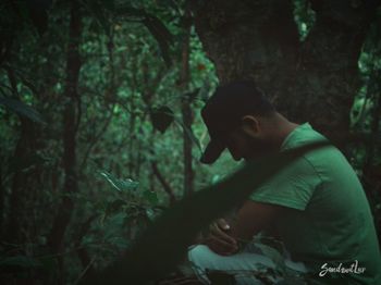 Young man looking away while sitting on tree in forest