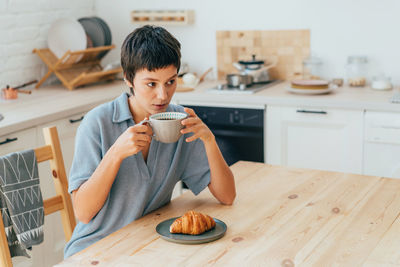 Young attractive european woman has coffee and croissant for breakfast while sitting in the kitchen