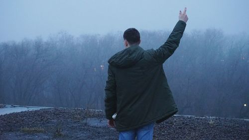 Rear view of man with arms raised against forest during foggy weather