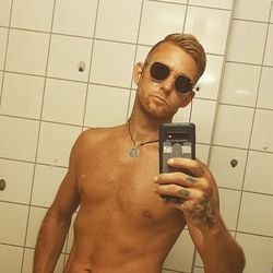 Portrait of man wearing sunglasses while taking selfie over mobile phone in bathroom