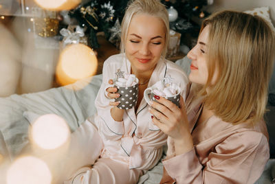 Two women at home drinking cocoa with marshmallows at the christmas tree. 