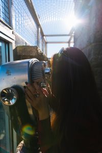 Side view of woman with long hair looking through coin-operated binoculars during sunny day