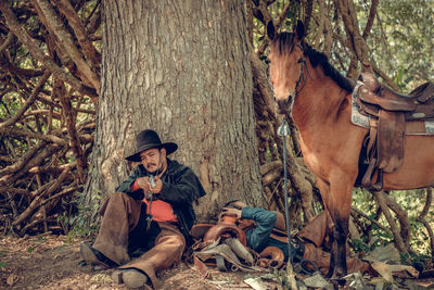 Man wearing hat aiming gun while sitting by tree in forest