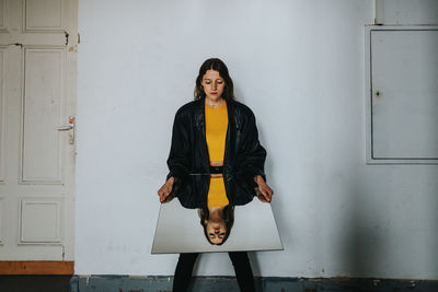 Young woman holding mirror while standing against wall