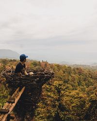 Young man looking at view while sitting on nest against cloudy sky