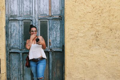 Portrait of young woman using mobile phone while standing at door