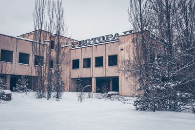 Bare trees on snow covered field by building against sky