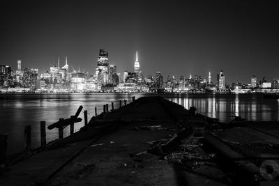 Damaged pier over river by illuminated cityscape against sky at night