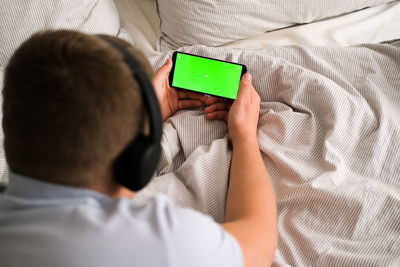 A man with headphones lies and holds a black mobile phone with a green screen upright