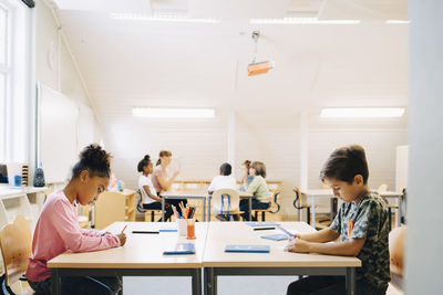 Boy and girl writing at desk while friends learning with student in background