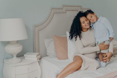 Cheerful mother embracing son sitting on bed at home