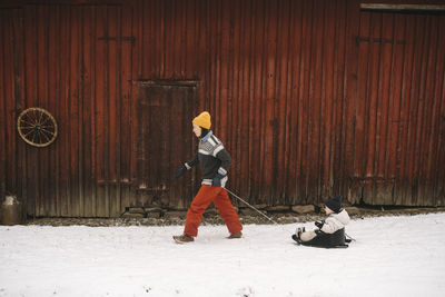 Mature woman pulling daughter on toboggan while walking by cottage during winter