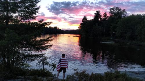 Rear view of man with american flag standing at lakeshore against sky during sunset