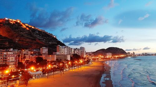Illuminated buildings by sea against sky at dusk