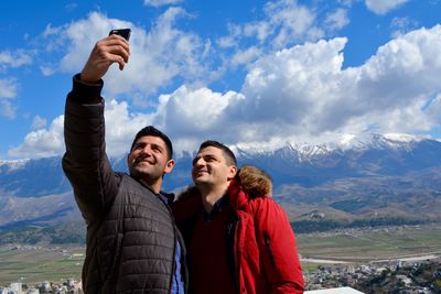 Friends taking selfie with phones with mountains in background against cloudy sky