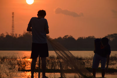 Rear view of silhouette mature men holding fishing net while standing by lake against sky during sunset