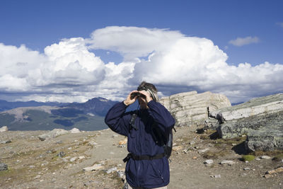 Hiker looking through binoculars while standing on mountain against sky