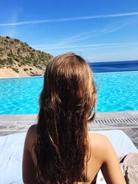 Rear view of woman with long hair looking at sea while sitting against blue sky during sunny day
