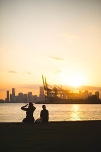 Silhouette people sitting by sea against sky during sunset